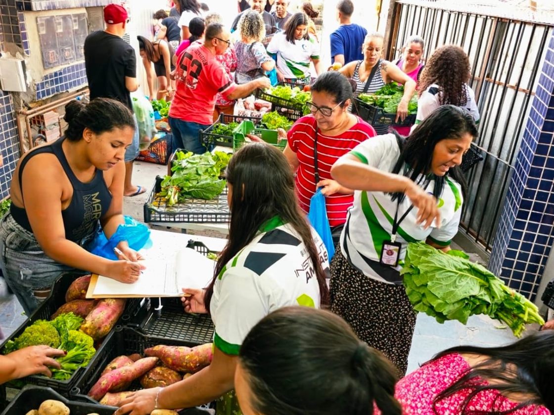 Distribuição de alimentos do Instituto: moradoras escolhem verduras enquanto uma voluntária faz o cadastro numa prancheta.